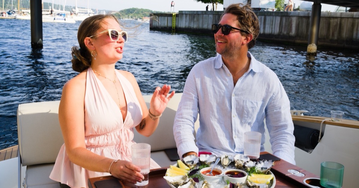 Two people on the aft deck of a private boat on the Oslofjord at sunset, the water copper-coloured toward the Oslo skyline
